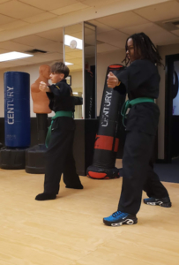 Two young teenage boys, one white and one Black, wearing black martial arts uniforms and green belts. They are standing in a martial arts stance and delivering a back fist in the air.
